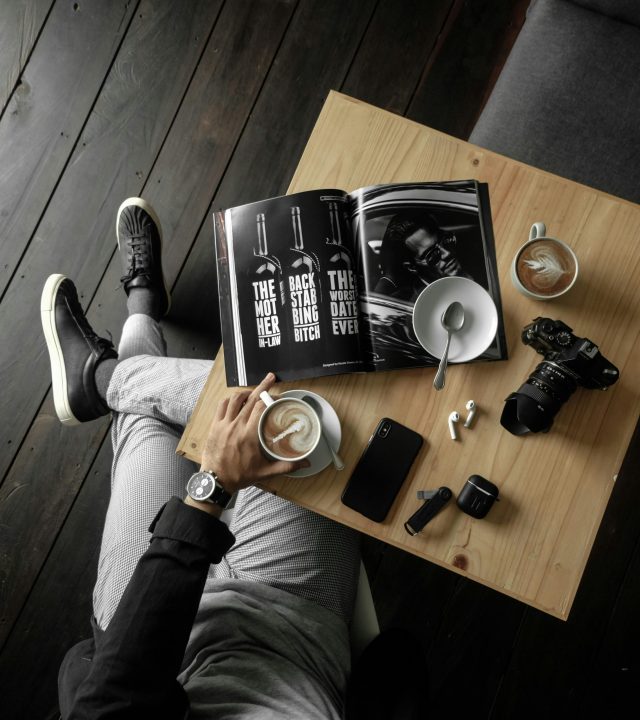 Top View Of A Table With Man Holding A Cup Of Latte With A Magazine, Camera, Cellphone And Other Personal Effects