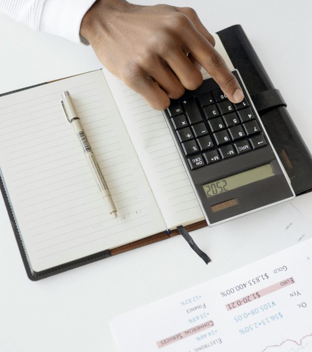 A hand using a calculator with financial documents and notebook, depicting an office setup for financial analysis.